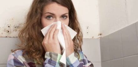 A woman is holding a cloth to her nose with both hands, and mold is visible on the wall behind her.